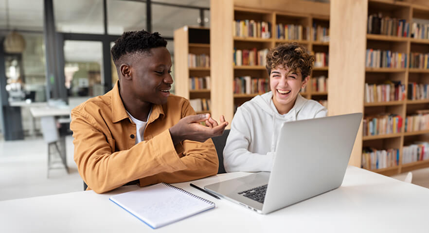 Zwei junge Menschen an einem Tisch mit einem laptop umgeben von Bücherregalen